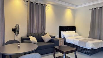 A hotel room with a bed, a sofa, a small table, a chair, and a standing fan.