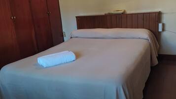 Habitación de hotel con dos camas, un armario de madera, un cuadro de árboles y una mesita de noche con una lámpara.