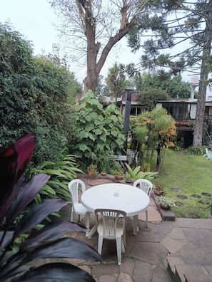 Un patio al aire libre con una mesa redonda y cuatro sillas, rodeado de abundante follaje y diversas plantas.