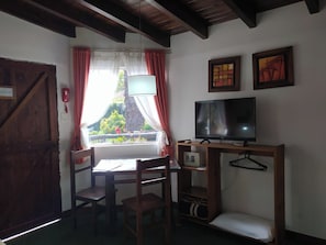 Una habitación con escritorio de madera, televisor sobre un mueble bajo, ventana con cortinas y una puerta.