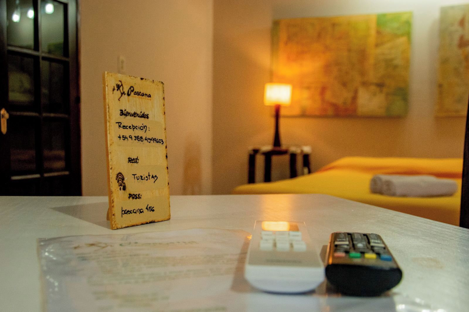 Habitación de hotel con un cartel de recepción, una cama, una lámpara y dos controles remotos sobre la mesa.