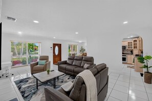Ein modernes Wohnzimmer mit Sofa, Sessel und Couchtisch. Durch einen Bogen ist eine Küche zu sehen, und eine Tür mit Fenster ist ebenfalls vorhanden.