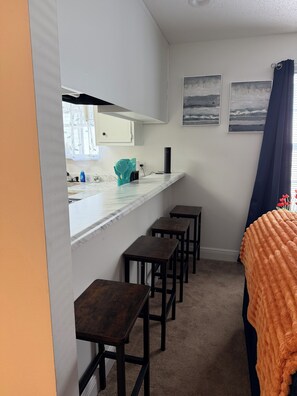 A compact kitchen with a white countertop, a black range hood, and a window with a curtain. There are three wooden stools and a dark-colored bed with an orange blanket. - Enjoyable 2-bedroom condo with AC in charming Pensacola (Pensacola)
