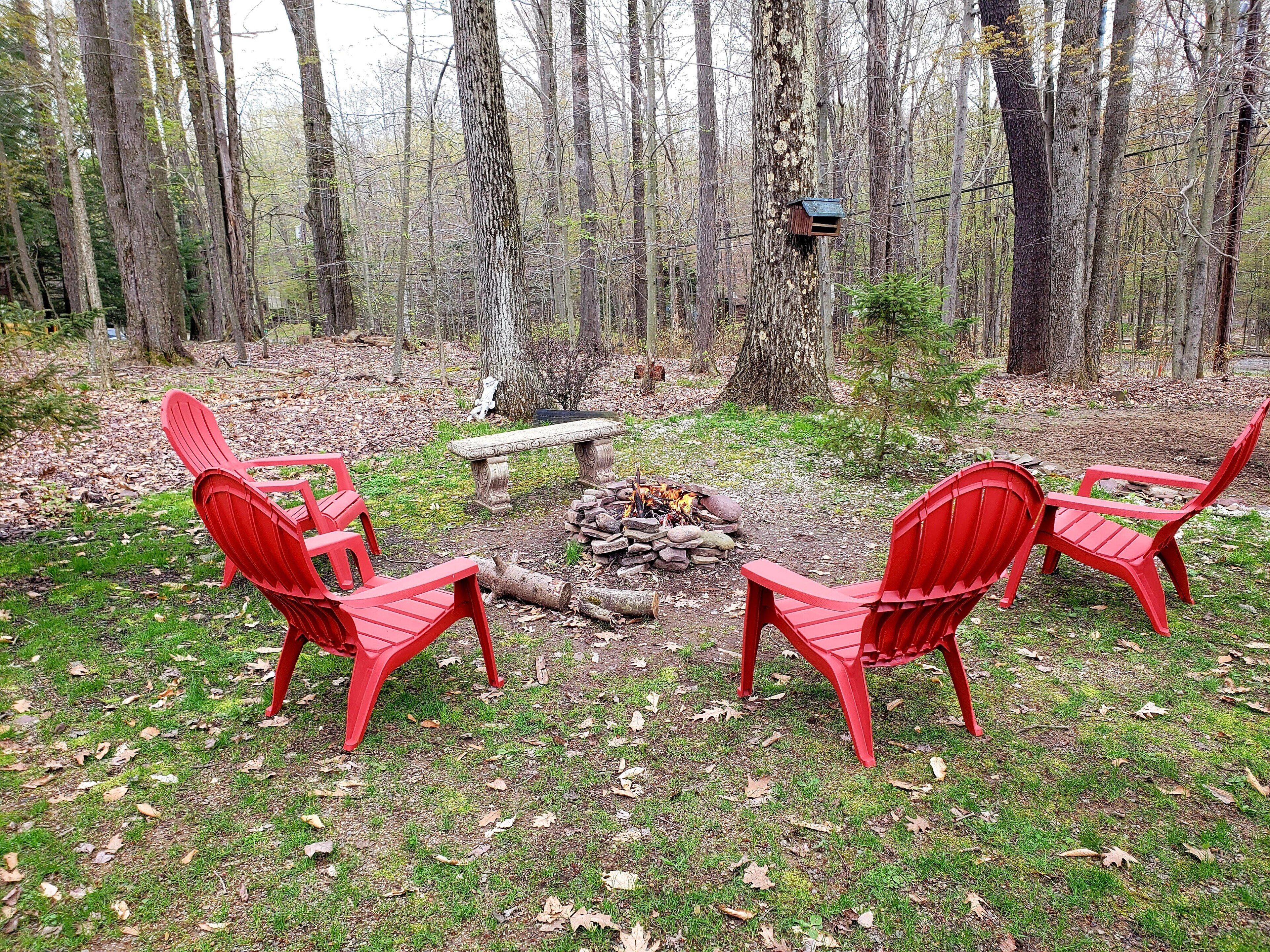A forest setting with a fire pit surrounded by red chairs.