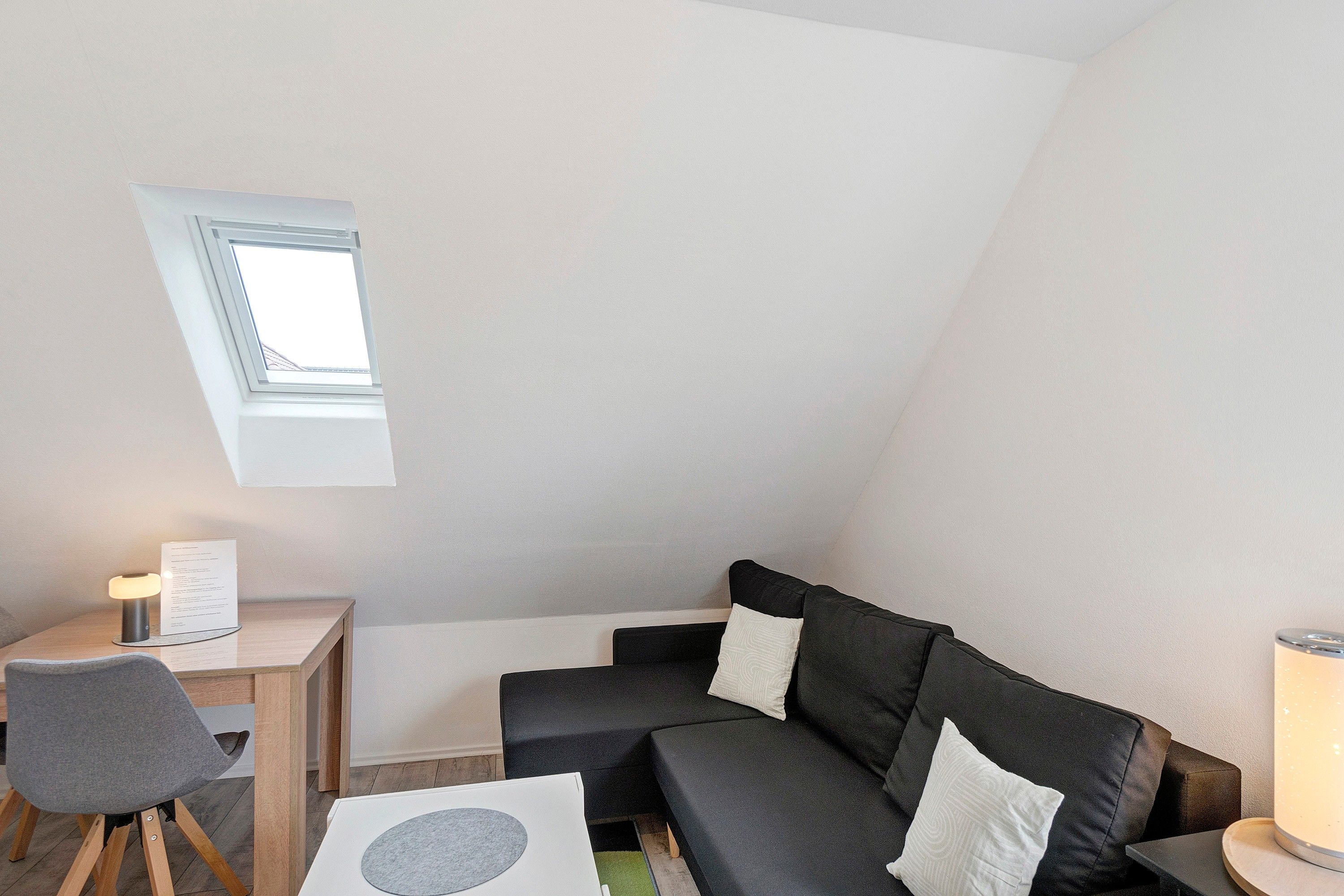 Et lille, hyggeligt værelse med skråvægge, et skrivebord med lampe, en stol, en sofa med puder og et vindue.
