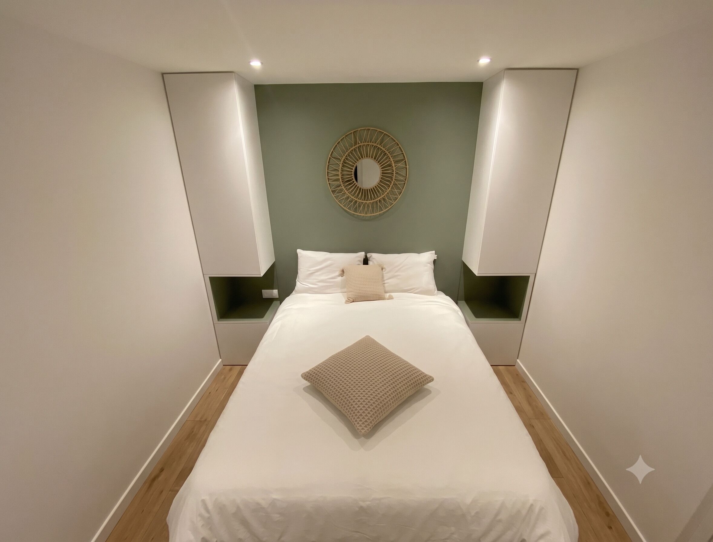 A bedroom with a bed centered in the room, flanked by nightstands, a wall-mounted circular mirror, and recessed ceiling lighting.