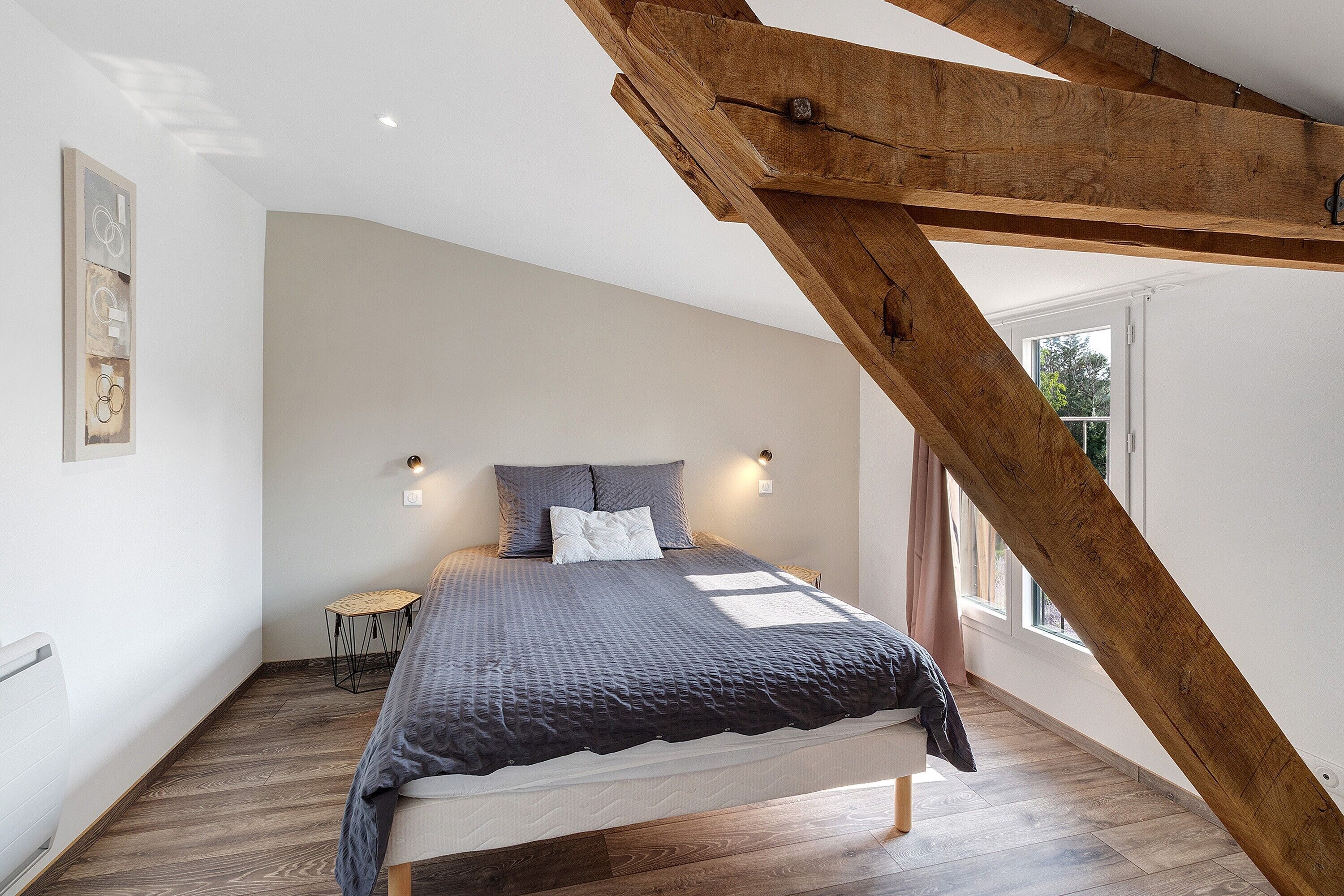 A bedroom with a sloped ceiling, a bed with a grey quilt, two bedside lamps, and a small table.