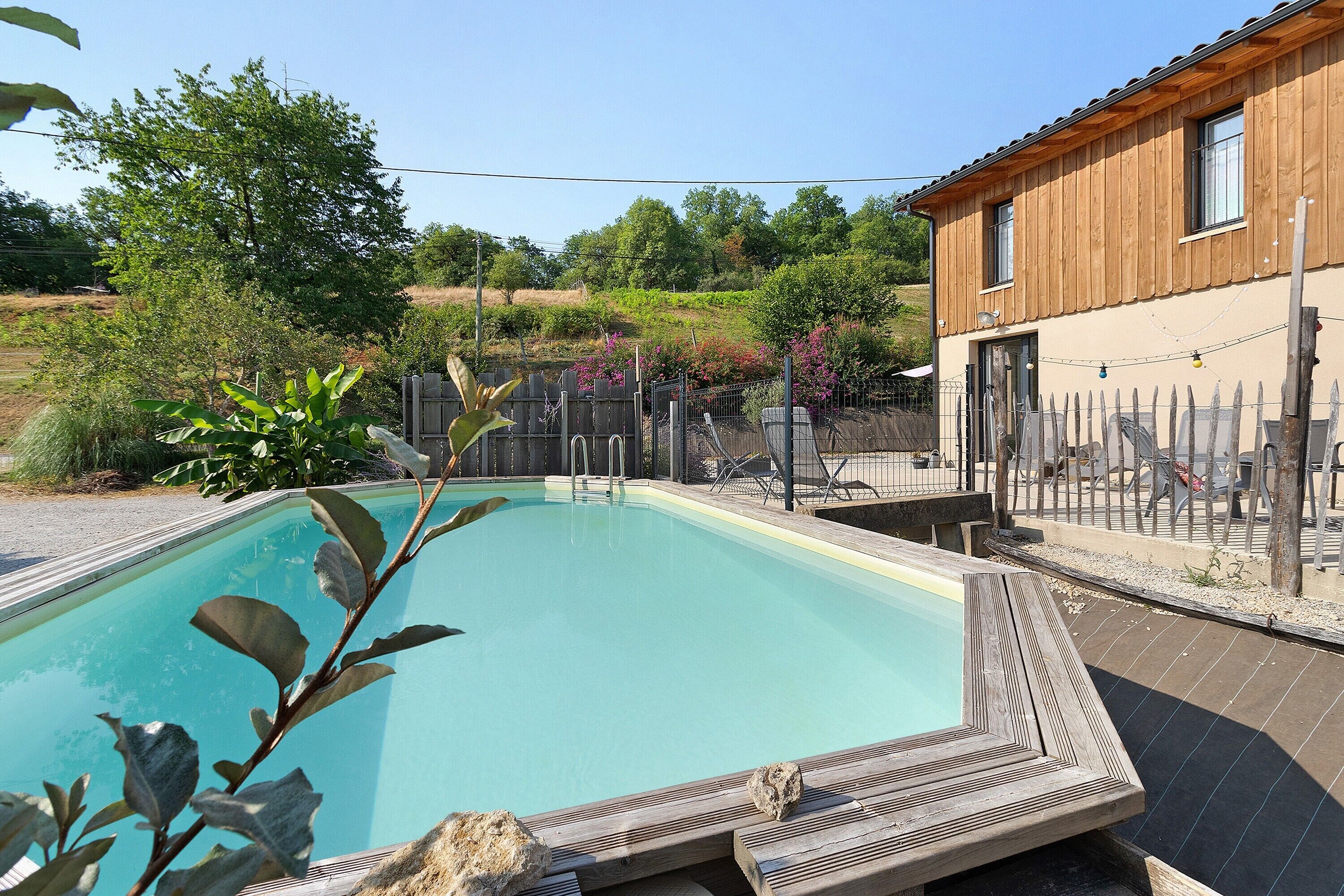 A swimming pool with a wooden deck, surrounded by a fence and greenery.