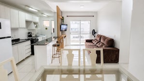 A modern kitchen with a central island, a dining area with a glass table, and a living space with a brown sofa. - Edifício Icaraí by LikeHome (Bombinhas)