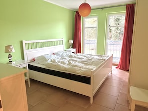 A bedroom with a white bed, green walls, a window with red curtains, and a small bedside table with a lamp. - Dünenresidenz Königin Augusta 2 in Binz (Ostseebad Binz)
