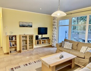 A living room with a sofa, coffee table, TV, and shelves with books and glassware. - Dünenresidenz Königin Augusta 2 in Binz (Ostseebad Binz)