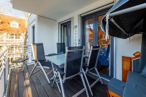 Uma varanda com mesa e cadeiras de vidro, um deck de madeira e vista para um edifício com uma janela.