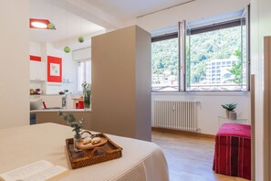 A modern bedroom with a large window, a bed, a small table with a tray, and a view of a building and trees. - Nineteen by Rent All Como (Como)