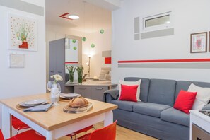 A modern living room with a grey sofa, red cushions, a wooden dining table with plates and a loaf of bread, and a kitchen area with cabinets and a hanging light fixture. - Nineteen by Rent All Como (Como)