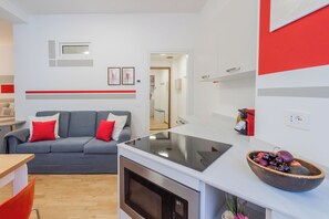 A compact living space with a kitchenette, a grey sofa with red cushions, and a small dining area. - Nineteen by Rent All Como (Como)