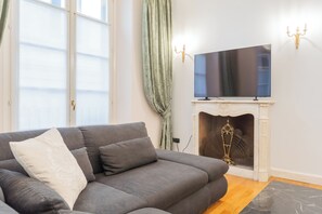 A living room with a grey sofa, a flat-screen TV mounted above a fireplace, and a window with sheer curtains. - Churchkey by Rent All Como (Como)