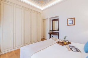 Ein Schlafzimmer mit großem Kleiderschrank, einem Bett mit weißer Bettdecke, einem Nachttisch mit Spiegel, einem gerahmten Bild und einer kleinen Pflanze.