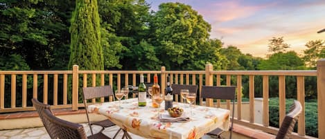 An outdoor patio with a table set for two, surrounded by wicker chairs, a bottle of wine, and a bowl of fruit.