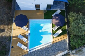 Vue aérienne d’une piscine entourée de chaises longues et de parasols, elle-même bordée d’une terrasse en bois et d’une zone herbeuse.