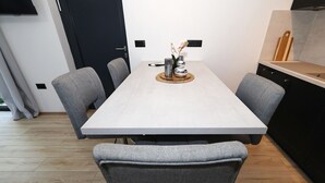 A dining area with a rectangular table, grey upholstered chairs, and a kitchen area with a black cabinet and a wooden cutting board. - Beachfront Almare Retreat (Sukošan)