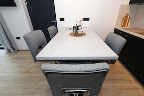 A dining area with a rectangular table, grey upholstered chairs, and a kitchen area with a black cabinet and a wooden cutting board. - Beachfront Almare Retreat (Sukošan)