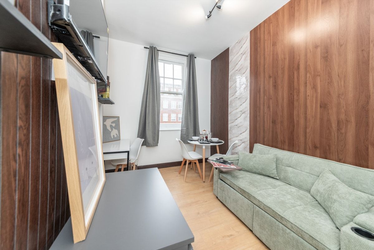 A modern living room with a sofa, a dining table with chairs, a small window with curtains, and a wall with a wood panel.