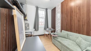 A modern living room with a sofa, a dining table with chairs, a small window with curtains, and a wall with a wood panel.