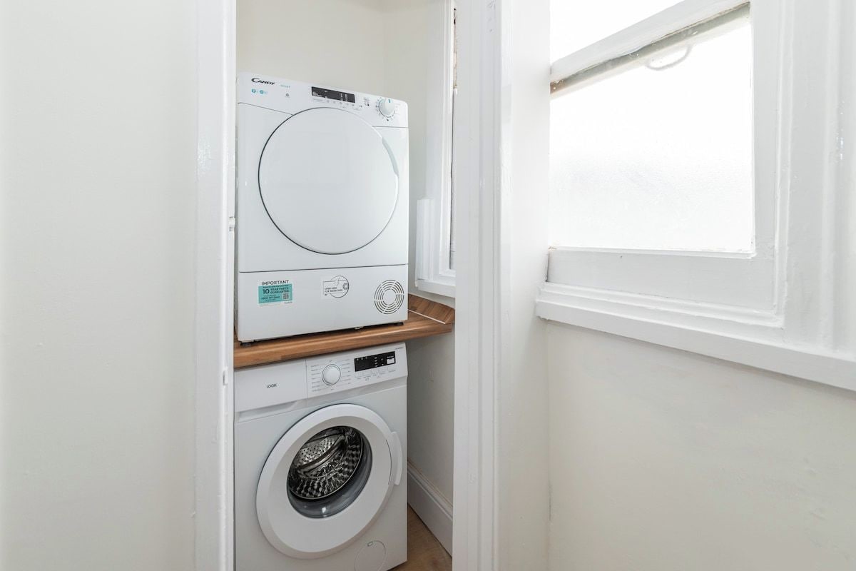 A compact laundry room with a front-loading washer and dryer, a wooden shelf, and a window providing natural light.