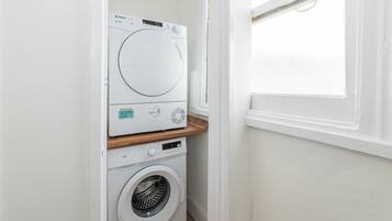A compact laundry room with a front-loading washer and dryer, a wooden shelf, and a window providing natural light.