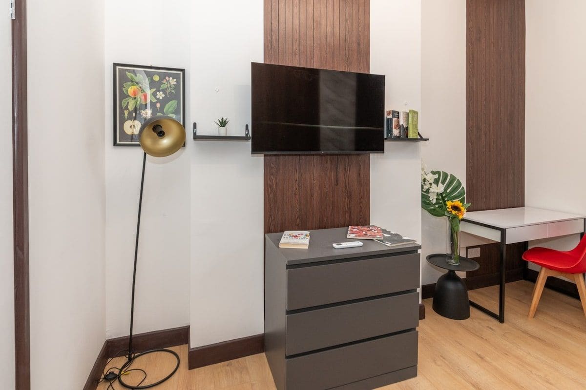 A modern room with a flat-screen TV mounted on the wall, a desk with a chair, a small plant, and a bookshelf.