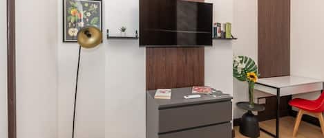 Een moderne kamer met een flatscreen televisie aan de muur, een bureau met stoel, een kleine plant en een boekenkast.