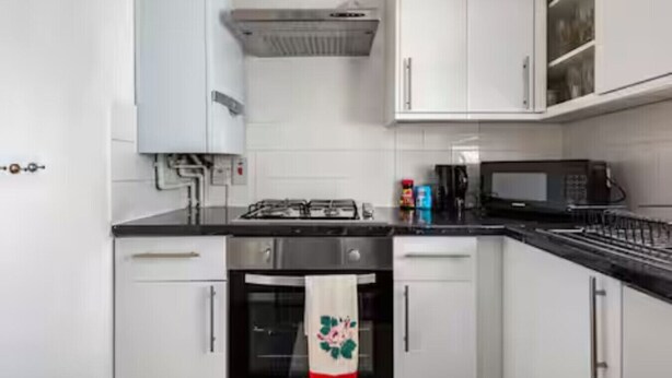A kitchen with white cabinets, a stove, a microwave, and a tiled backsplash. - Bright Maida Vale Flat near Shops- Transport Links (London)