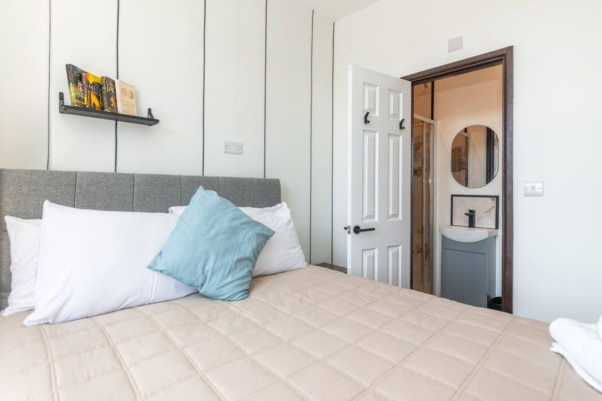 Un dormitorio con cama, almohadas, una mesita de noche con libros, una puerta y un espejo.