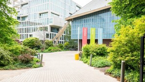 A modern building with a glass facade, a paved walkway, and greenery. - Stylish New Studio in Kensington with AC (Greater London)