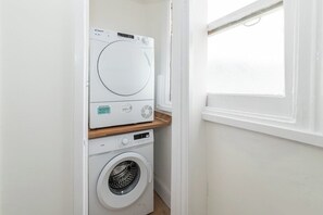 A compact laundry room with a front-loading washer and dryer, a wooden shelf, and a window providing natural light. - Lumen Kensington - Brand New - Airconditioned - W8 (Greater London)