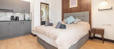 A bedroom with a large bed, a bedside table, a wooden headboard, and a view of the kitchen.