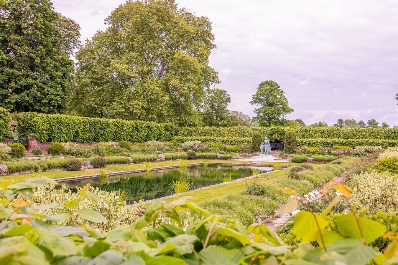 A formal garden with a central pond, statues, and neatly trimmed hedges.