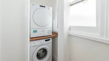 A compact laundry room with a front-loading washing machine and dryer, a wooden shelf, and a window.