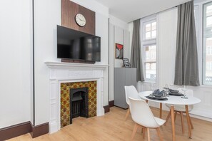 Un salon moderne doté d’une cheminée, d’un téléviseur à écran plat fixé au-dessus, d’un coin repas avec une table et des chaises, et d’une grande fenêtre agrémentée de rideaux.