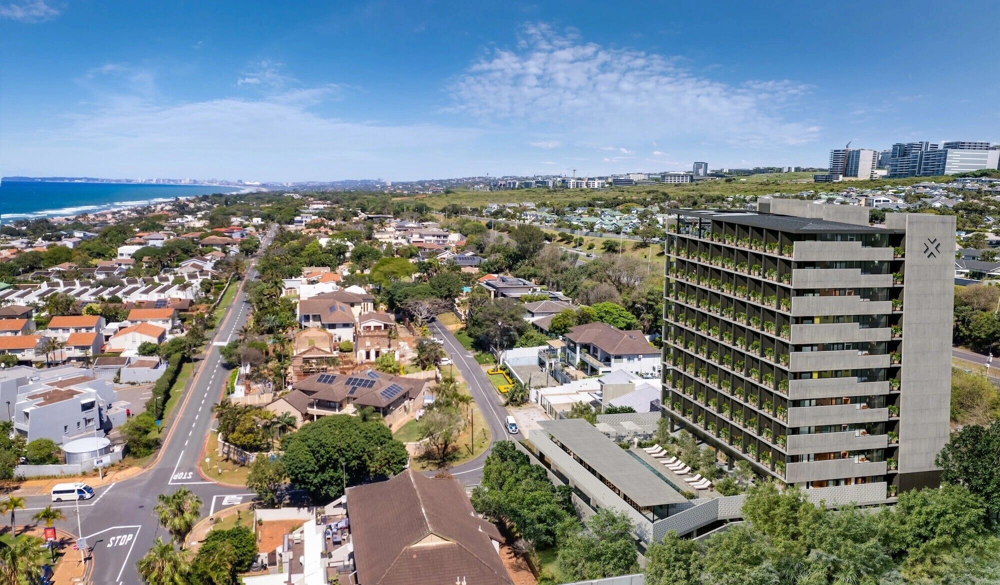 A high-rise building with multiple balconies, surrounded by a residential area with houses and a coastal view.