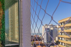 A chain-link fence with a view of high-rise buildings and a body of water in the background. - Apt with Balcony & Parking 300m | Vila Tupi Beach (Praia Grande)