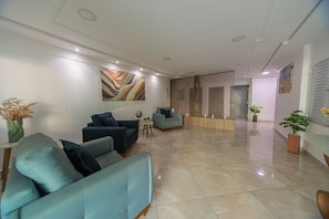 A modern lobby with seating areas, a reception desk, and a large abstract painting on the wall. - Apt with Balcony & Parking 300m | Vila Tupi Beach (Praia Grande)