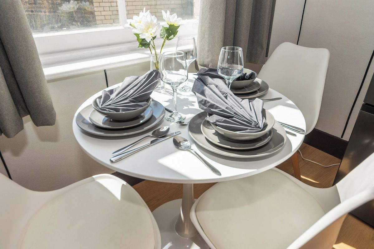 A dining table set for two with white chairs, a vase with white flowers, and a window with curtains.