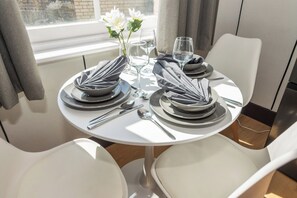 A dining table set for two with white chairs, a vase with white flowers, and a window with curtains. - Halcyon Kensington 1 Bedroom - Airconditioned (Greater London)