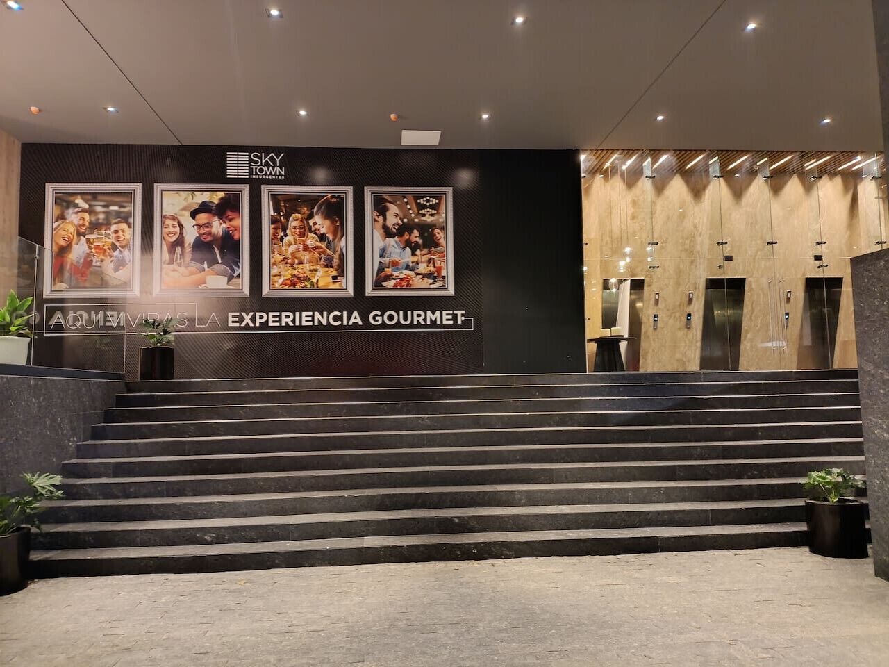 Eine Hotelhalle mit einer Treppe, die zu einem Aufzug führt, einem Empfangsbereich und einer Wand mit gerahmten Bildern.