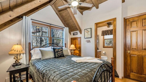 Une chambre avec un grand lit, un plafond en bois, une fenêtre donnant sur les arbres, une porte et une table de chevet avec une lampe.