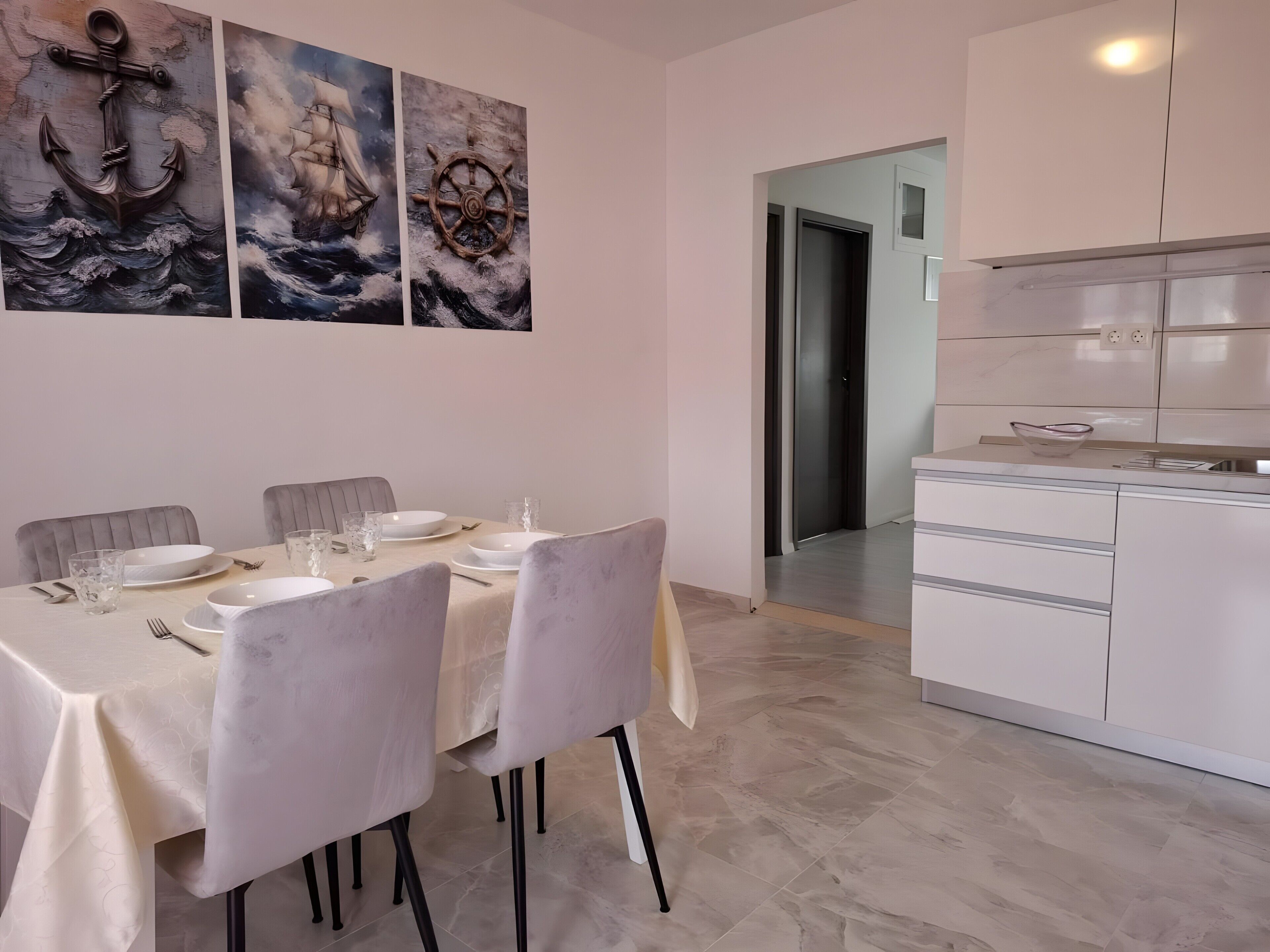 Un coin repas avec une table dressée pour quatre personnes, une cuisine avec des meubles blancs et un couloir menant à une autre pièce.