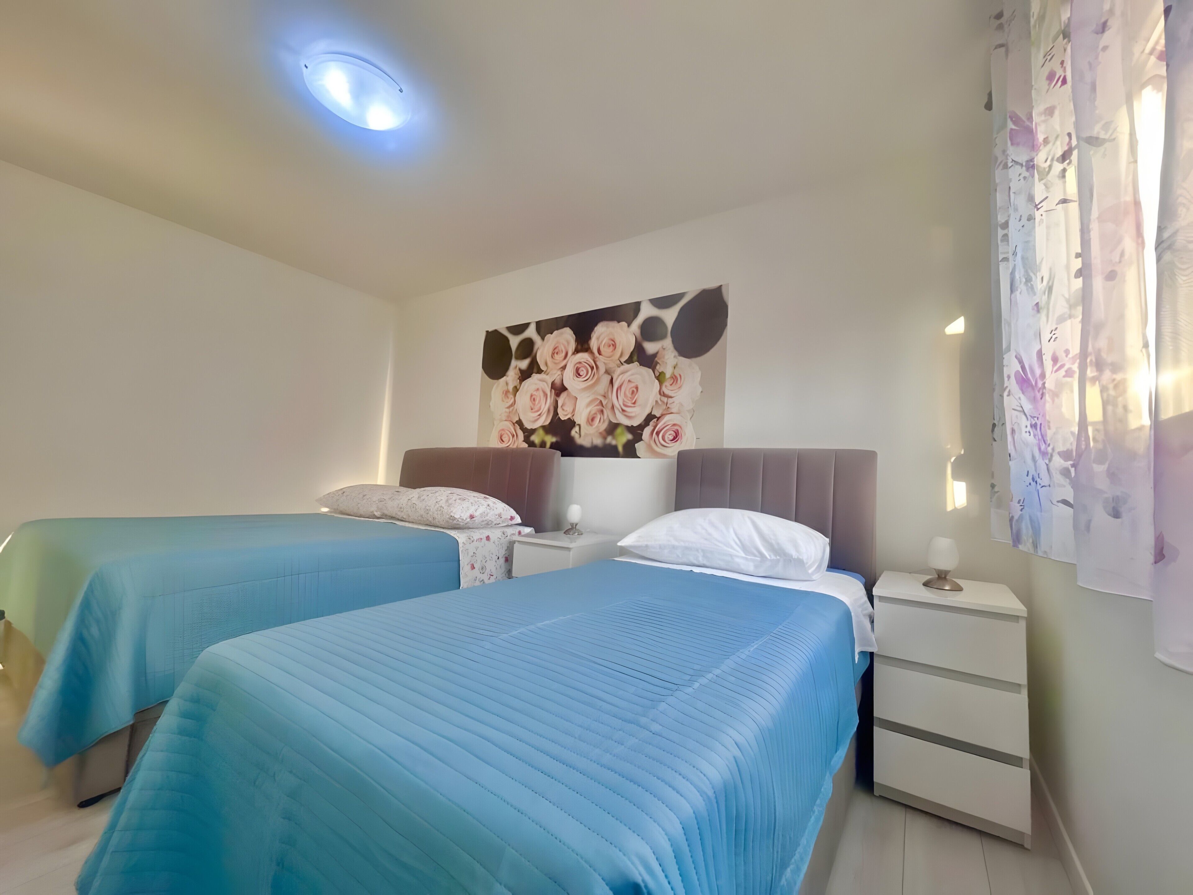 A hotel room with two beds, a nightstand, a lamp, a floral painting, and a curtain.