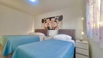 A hotel room with two beds, a nightstand, a lamp, a floral painting, and a curtain.