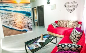 Ein Wohnzimmer mit einem roten Sofa, gemusterten Kissen, einem Glastisch mit Zeitschriften und einem Wandbild mit Strandmotiv.
